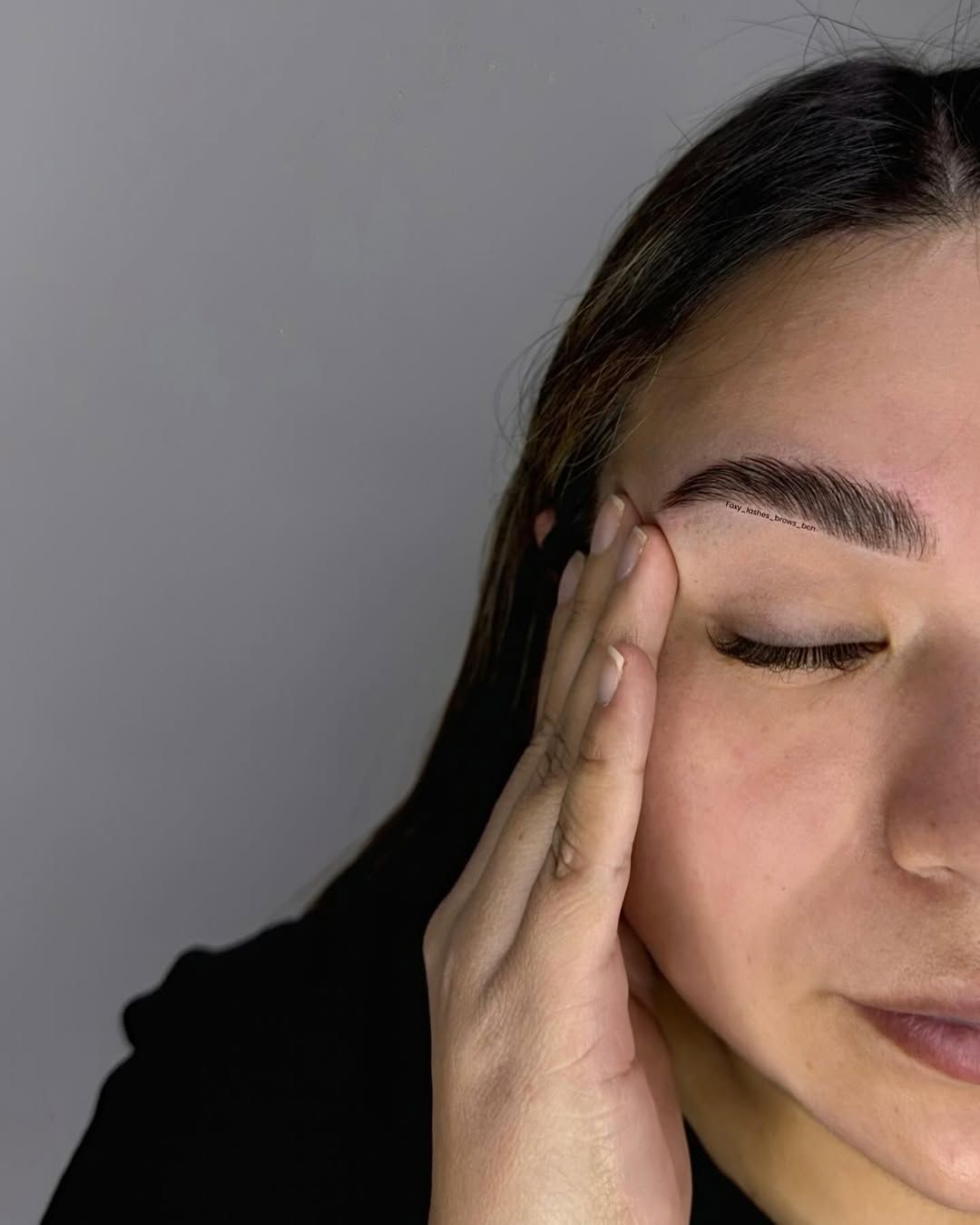 Primer plano de una persona tocándose la sien con los ojos cerrados sobre un fondo gris liso.