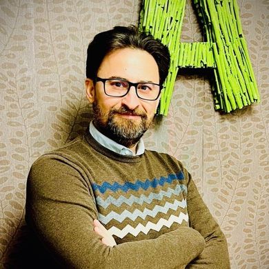 Un hombre con gafas y barba está de pie con los brazos cruzados delante de una letra a verde.
