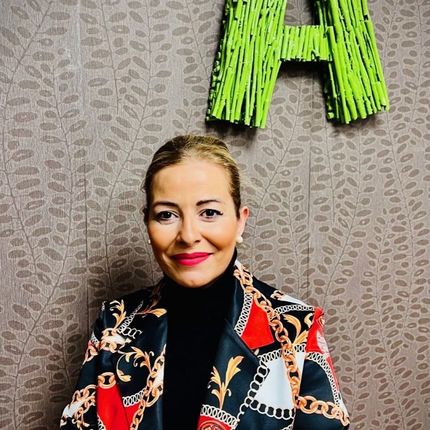 Una mujer con una chaqueta colorida sonríe delante de una letra a verde.