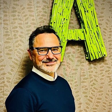 Un hombre que lleva gafas y un suéter azul está parado frente a una letra a verde.