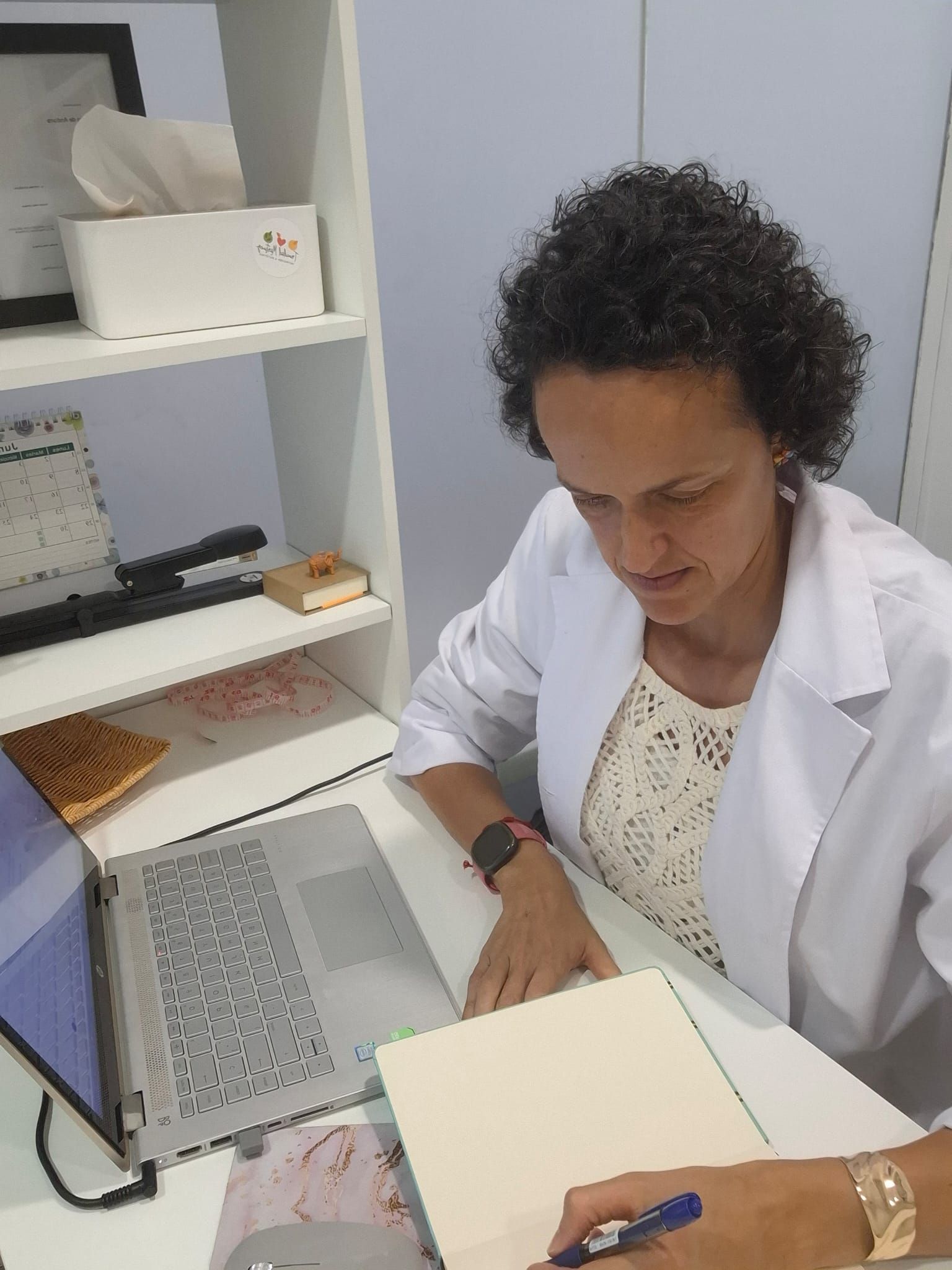 Persona con bata blanca trabajando en un escritorio con una computadora portátil y un cuaderno en una pequeña oficina.