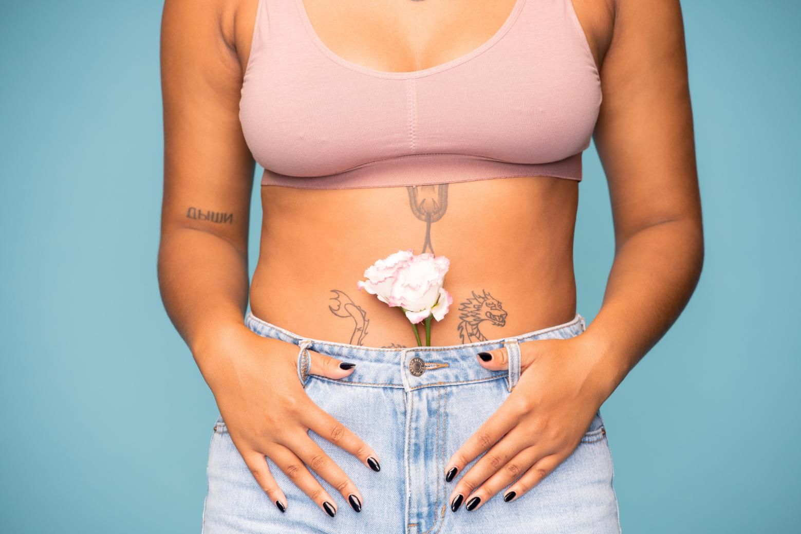Torso de una persona con una blusa rosa y jeans, con una flor blanca en la parte inferior del abdomen sobre un fondo azul.