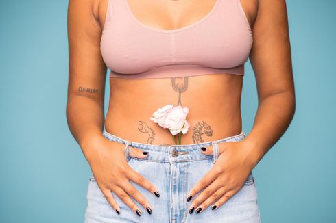 Torso de una persona con una blusa rosa y jeans, con una flor blanca en la parte inferior del abdomen sobre un fondo azul.