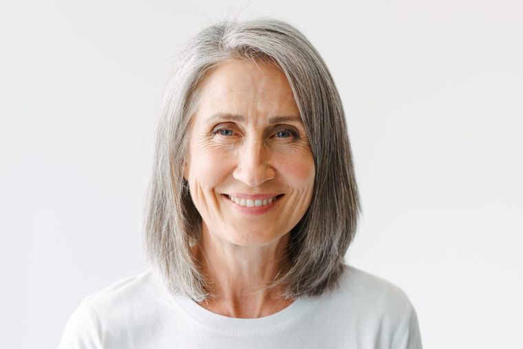 Mujer sonriente con cabello gris y camisa blanca sobre un fondo claro liso.