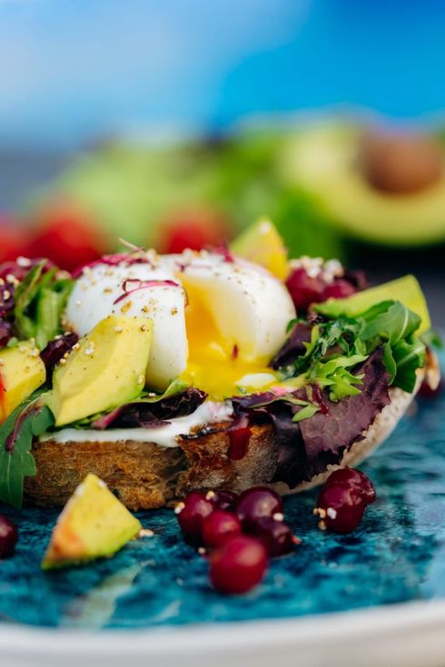 Tostada de aguacate con huevo escalfado y verduras, servida en un plato azul con frutos rojos.