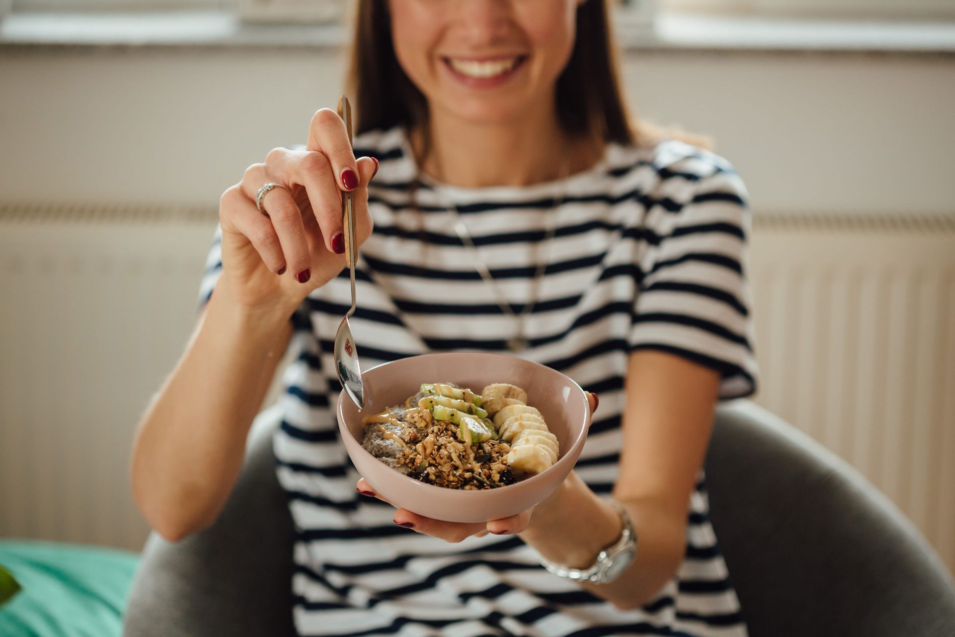 Mujer con camisa de rayas, sosteniendo un tazón de comida y un tenedor, sonriendo en un sofá.
