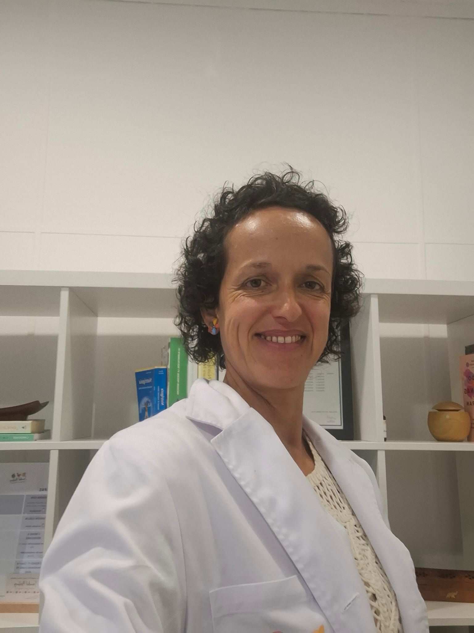 Persona con bata blanca de laboratorio sonriendo en una oficina o clínica, de pie frente a unas estanterías.