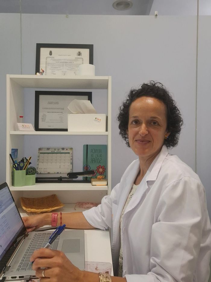 Persona con bata blanca de laboratorio trabajando en un ordenador portátil en una oficina con certificados en una estantería.