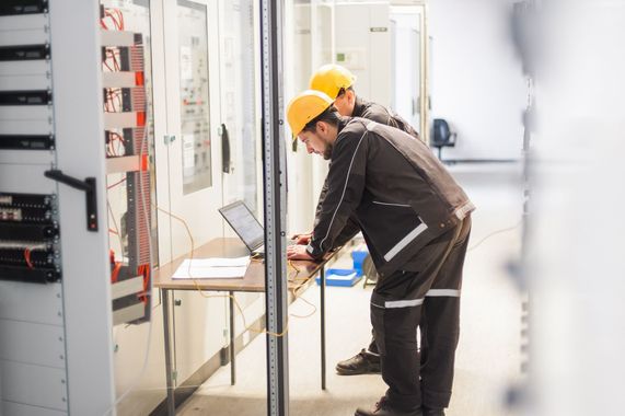 Un ouvrier portant un casque jaune inspecte du matériel électrique dans une salle des machines lumineuse.
