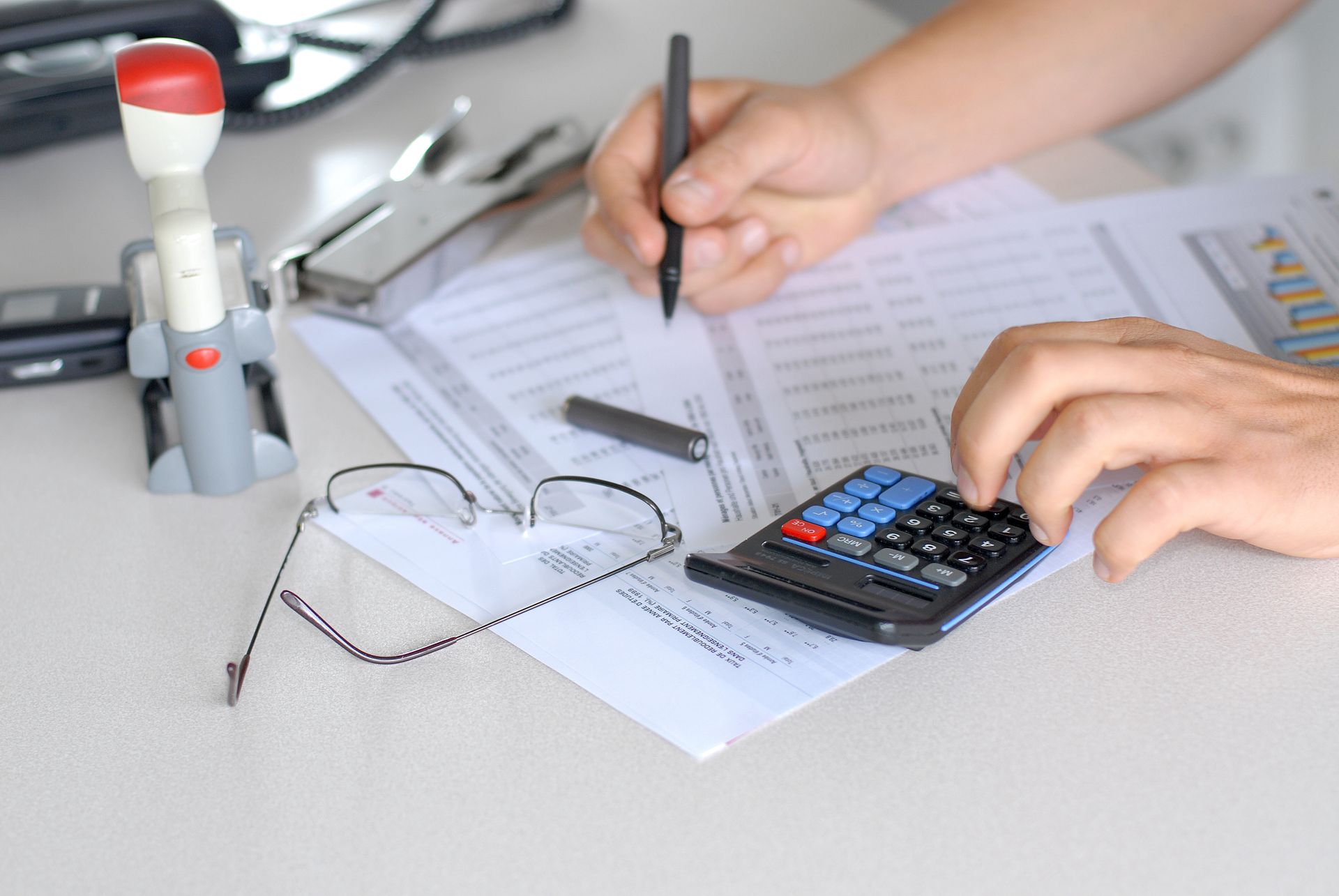 Des mains manipulant une calculatrice et écrivant sur des documents, à côté de lunettes et de fournitures de bureau.