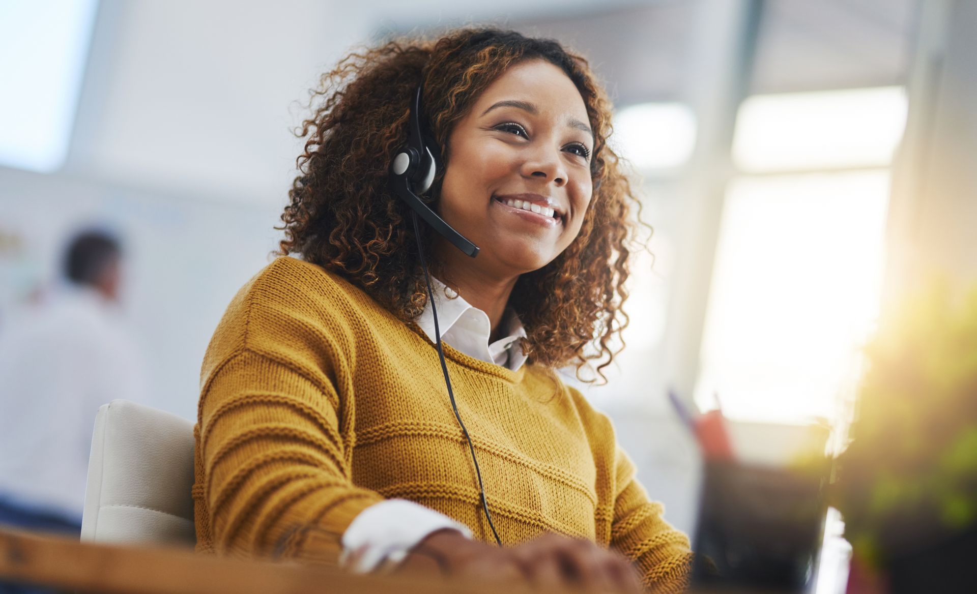 Une femme souriante portant un casque audio et un pull moutarde est assise dans un bureau lumineux.