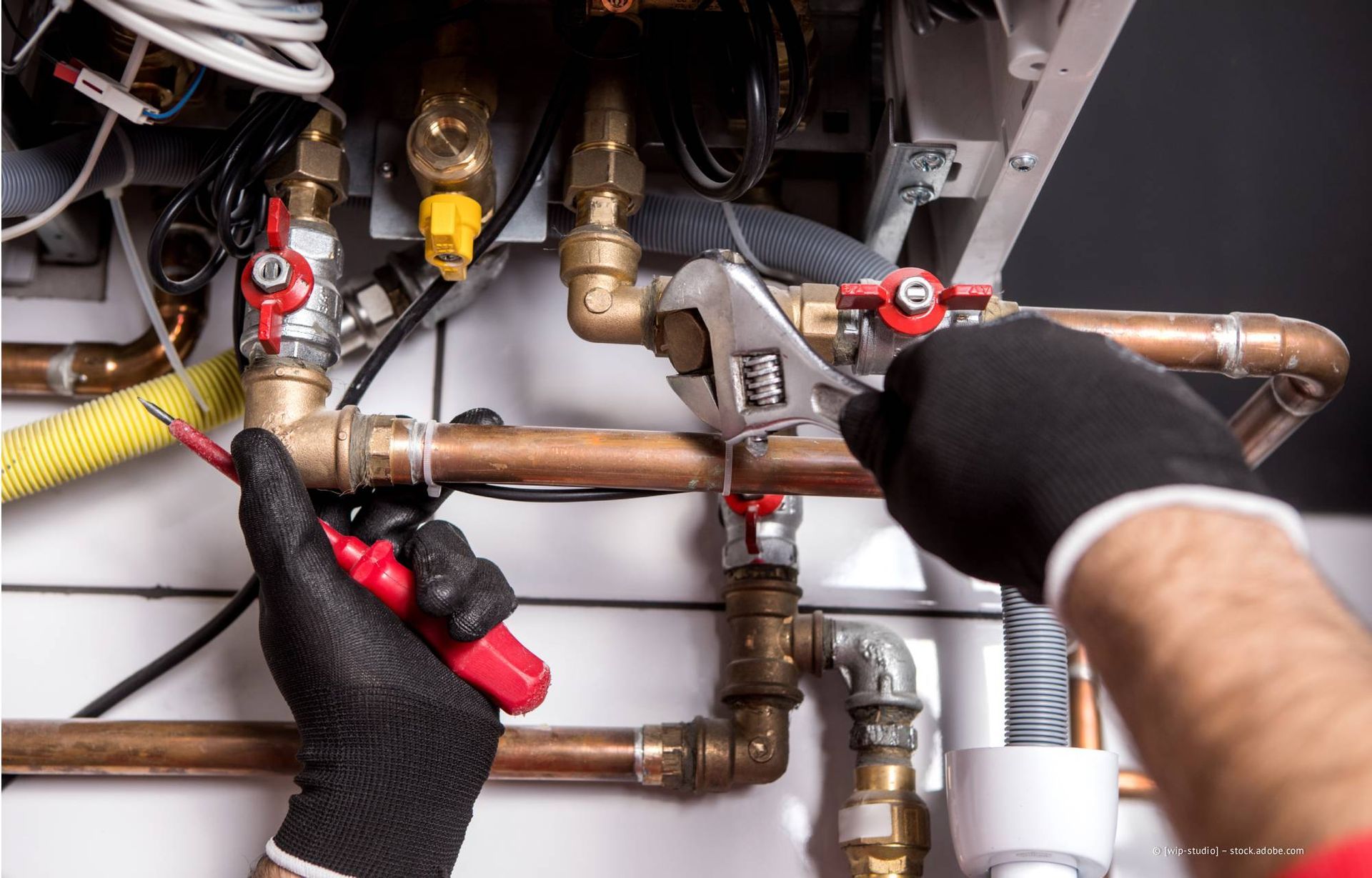 Ein Klempner, der mit Schraubenschlüssel und Zange an Kupferrohren in der Nähe eines Heizkessels arbeitet, trägt schwarze Handschuhe.