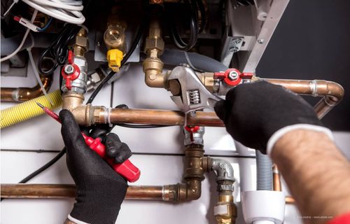 Ein Klempner, der mit Schraubenschlüssel und Zange an Kupferrohren in der Nähe eines Heizkessels arbeitet, trägt schwarze Handschuhe.