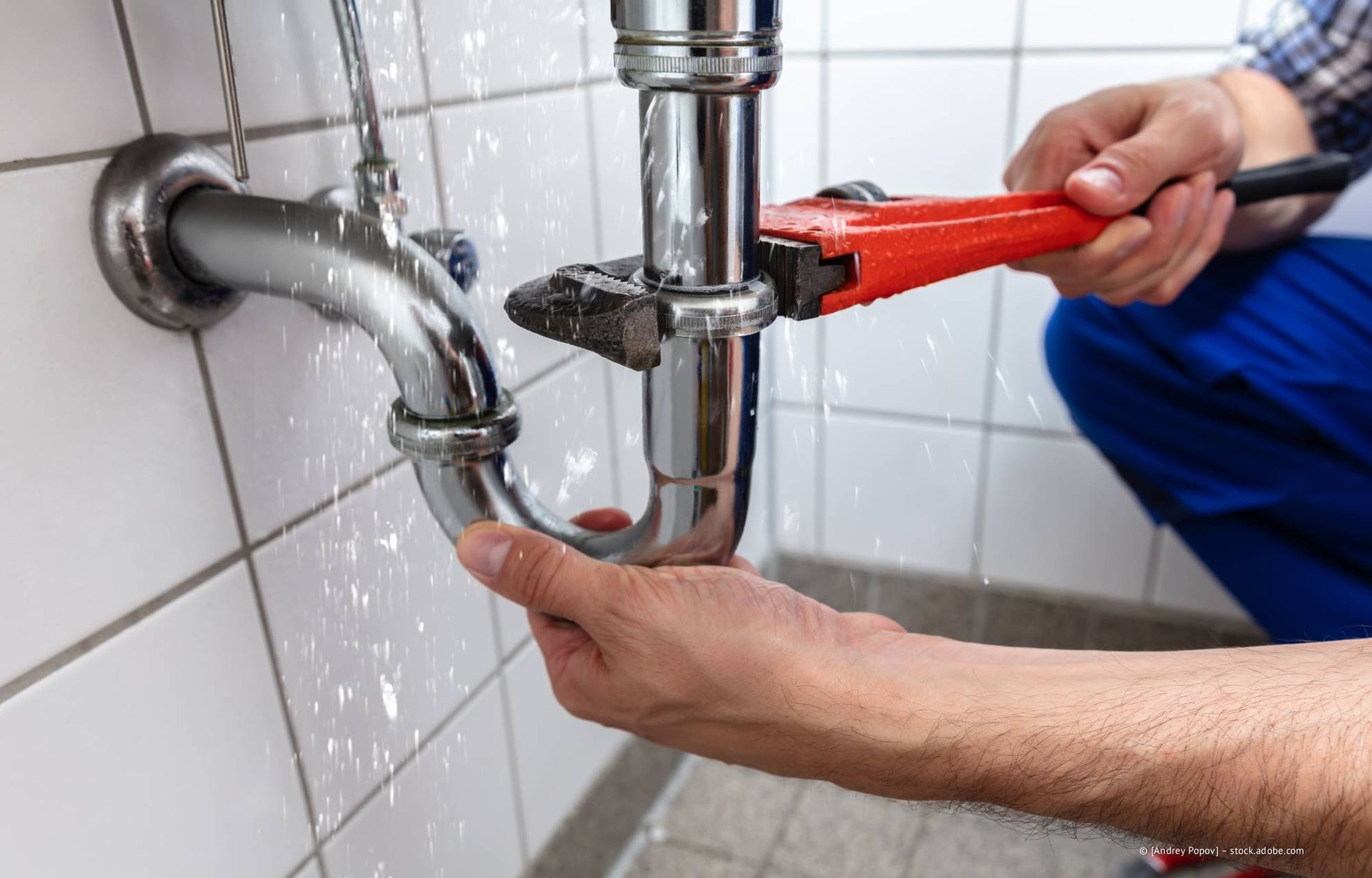 Ein Klempner benutzt einen Schraubenschlüssel, um ein undichtes Waschbeckenrohr in einem gefliesten Badezimmer zu reparieren.