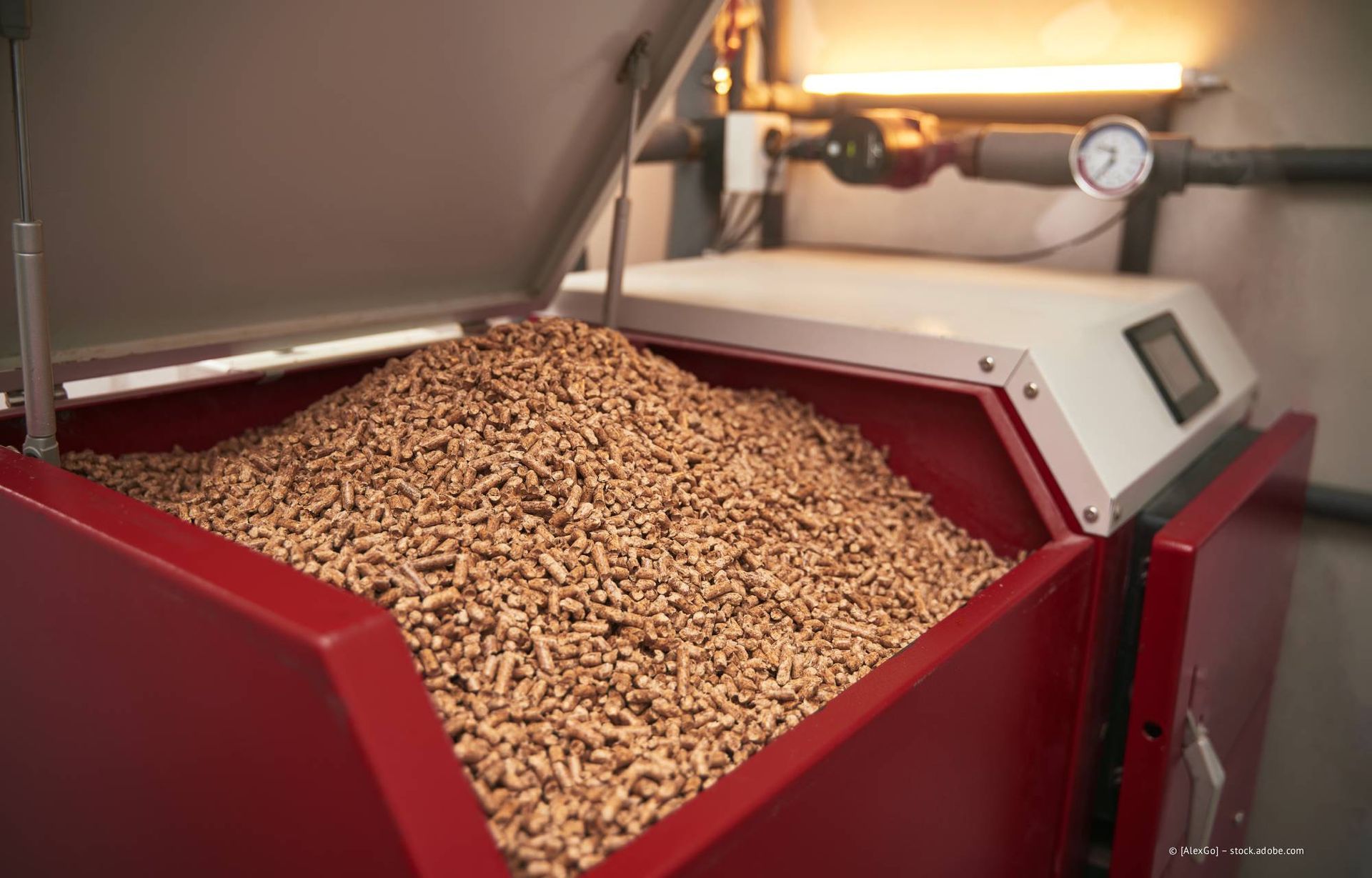 Rote Holzpellet-Heizanlage, Deckel geöffnet, gefüllt mit braunen Pellets; Rohre und Manometer im Hintergrund sichtbar.