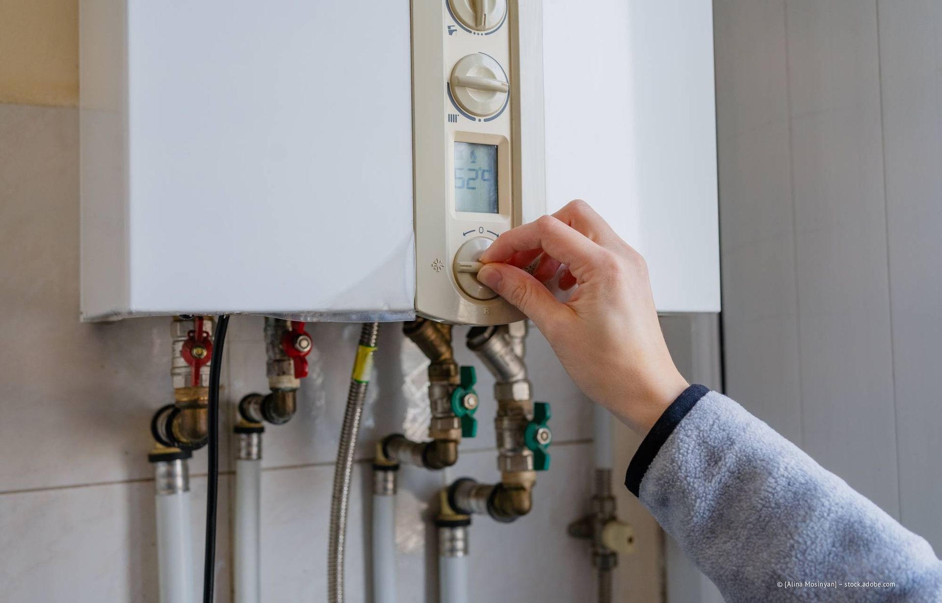 Eine Hand dreht an einem Drehknopf an einem weißen, an der Wand montierten Heizkessel in der Nähe von Rohren und Ventilen.