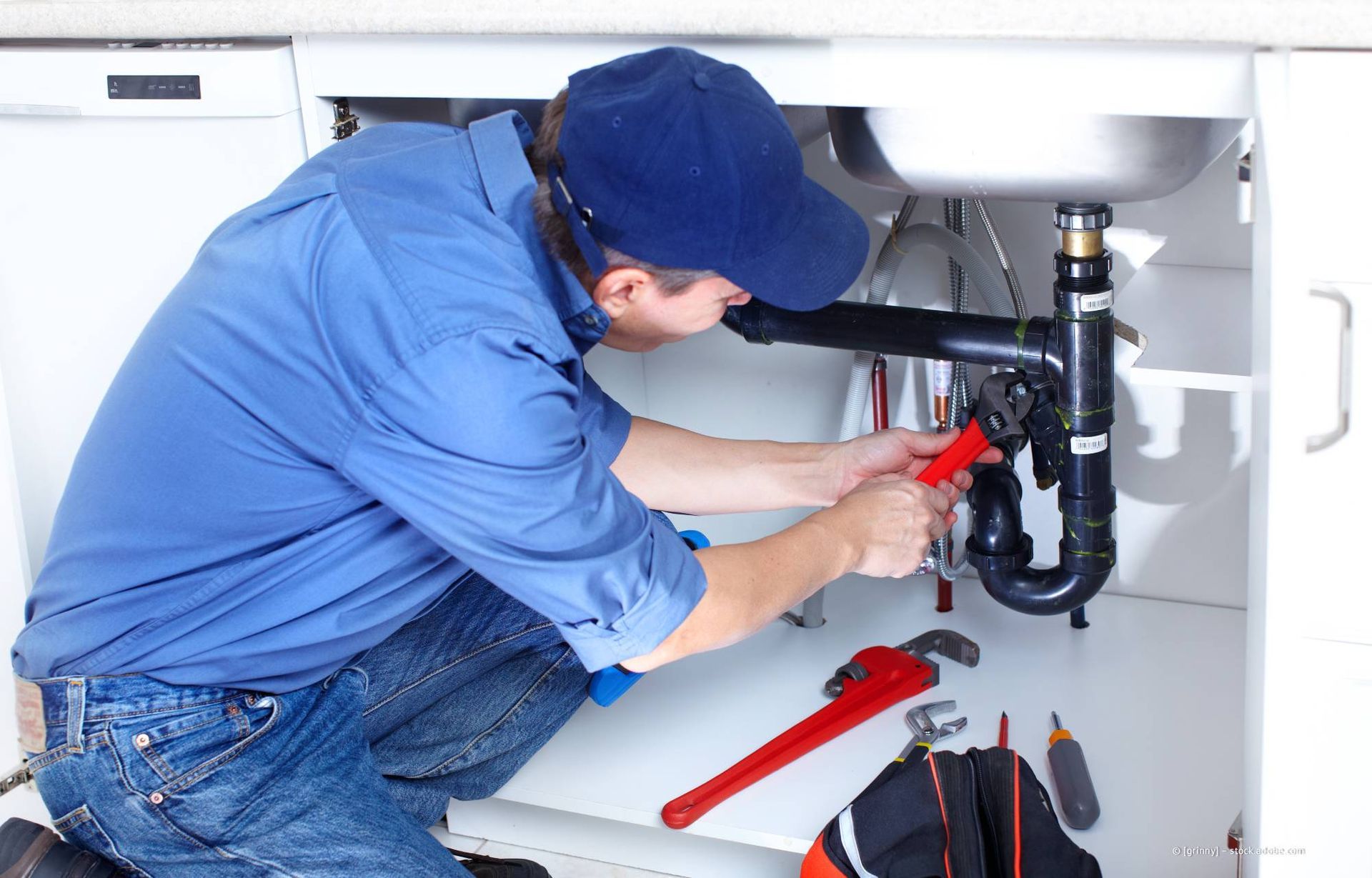 Ein Klempner in blauem Hemd und Kappe arbeitet unter einer Küchenspüle und benutzt dabei Werkzeuge.