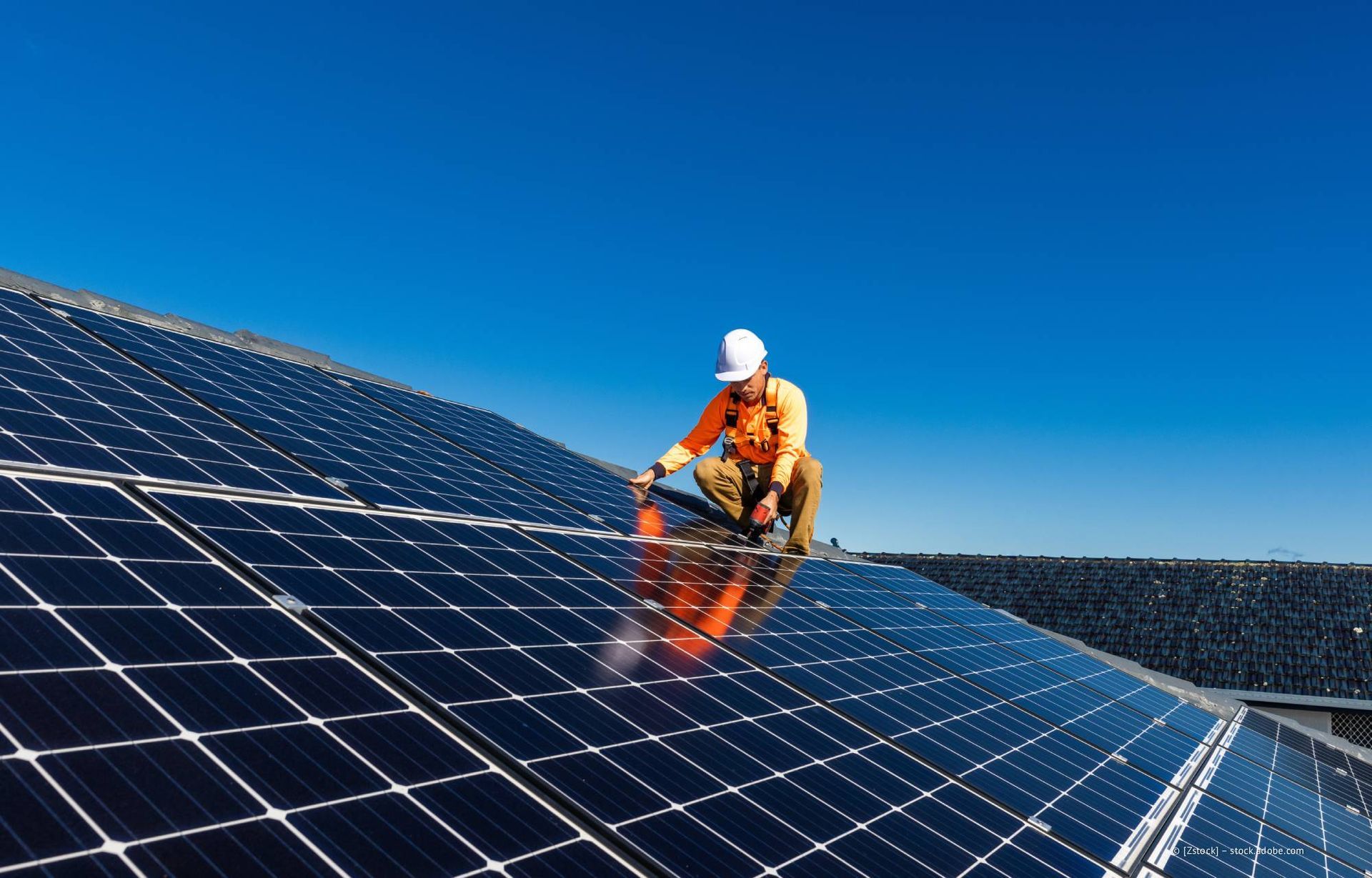 Ein Arbeiter in orangefarbener Schutzkleidung und weißem Schutzhelm installiert auf einem Dach unter strahlend blauem Himmel Solarpaneele.
