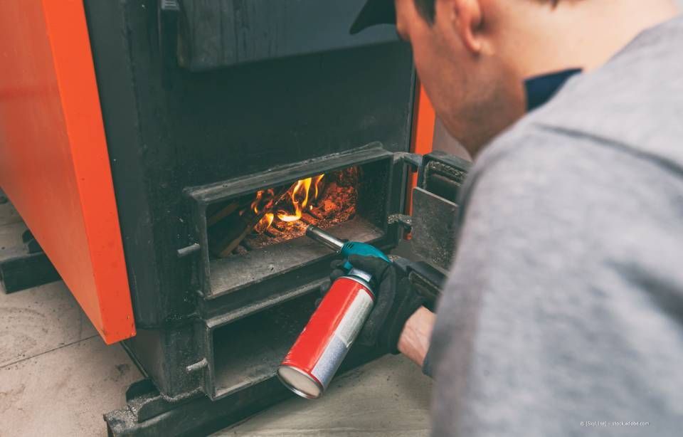 Person entzündet Holz in einem Ofen mit einer Fackel, orange und schwarze Farben, Innenaufnahme.