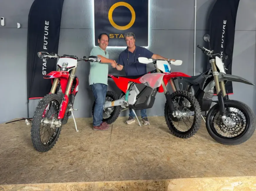 Dos personas posando con tres motos de motocross en una sala de exposición, entre ellas motos rojas, blancas y negras.