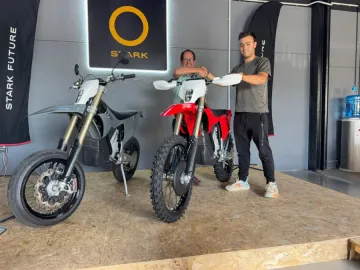 Dos personas de pie junto a dos motos de motocross en una sala de exposición, una negra y otra roja.