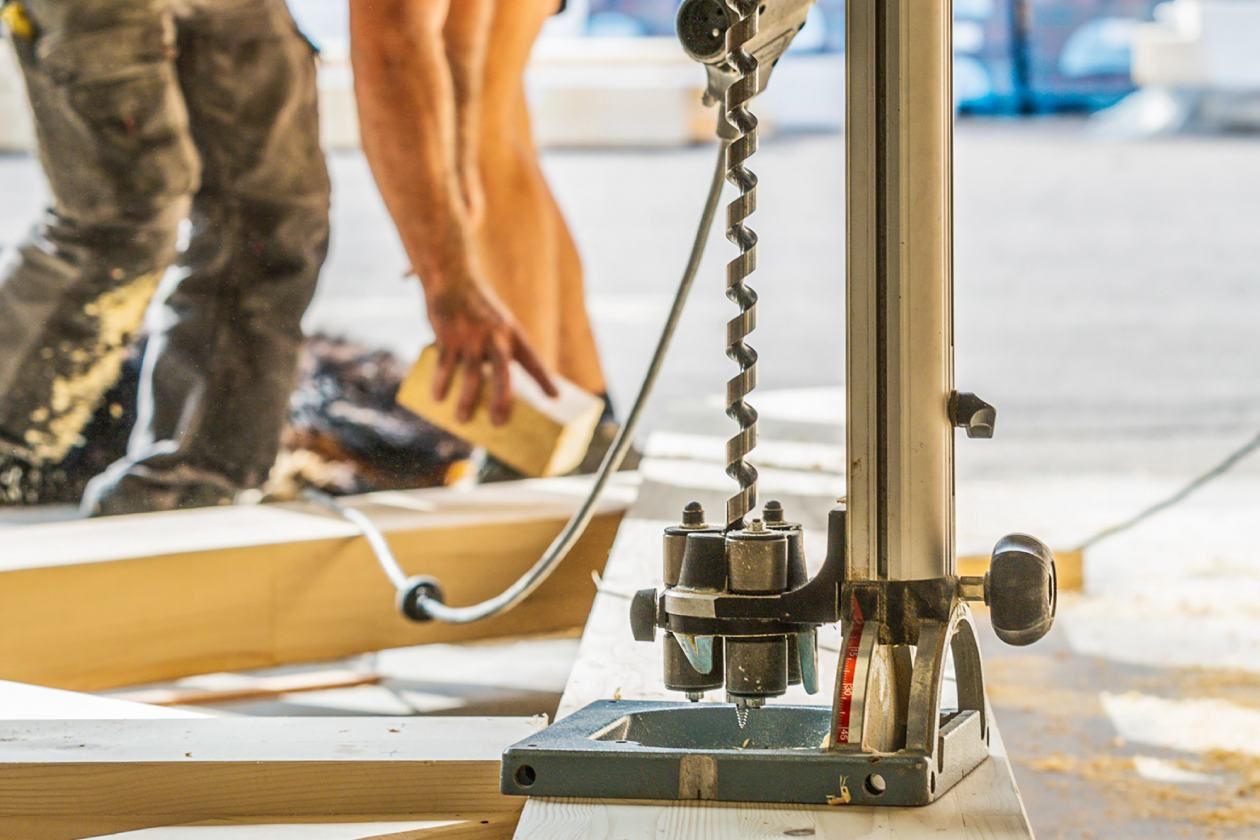 Gros plan sur une perceuse à colonne avec un charpentier qui manipule des pièces de bois en arrière-plan.