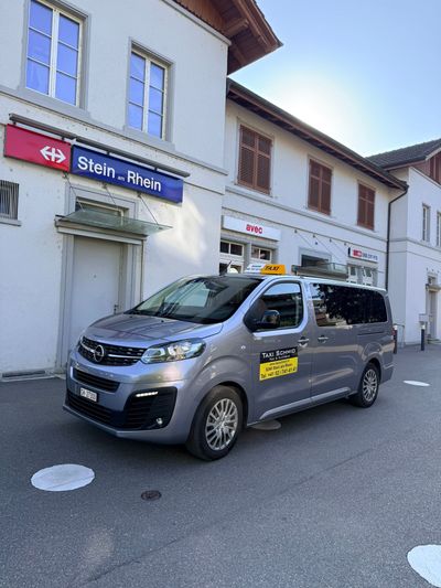 Ein weisser Lieferwagen parkt in einer sonnigen Strasse, an der Seite steht „Taxi Schmid“.
