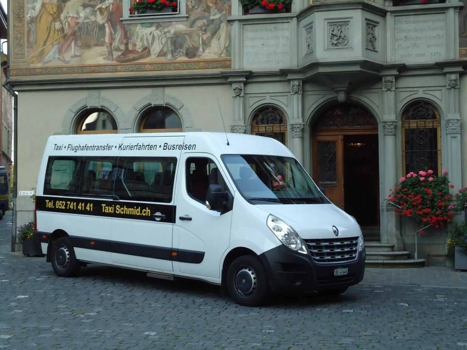 Ein weisser Lieferwagen parkt vor einem Gebäude mit Bogeneingang. Auf der Seite steht: „Taxi Schmid“.