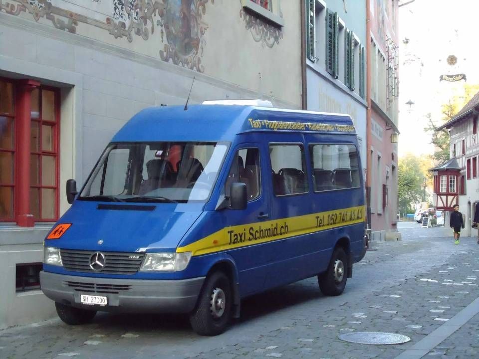 Ein blauer Mercedes-Benz-Transporter mit der Aufschrift „Taxi Schwyz“ parkt auf einer Kopfsteinpflasterstrasse. Eine Person sitzt am Steuer.