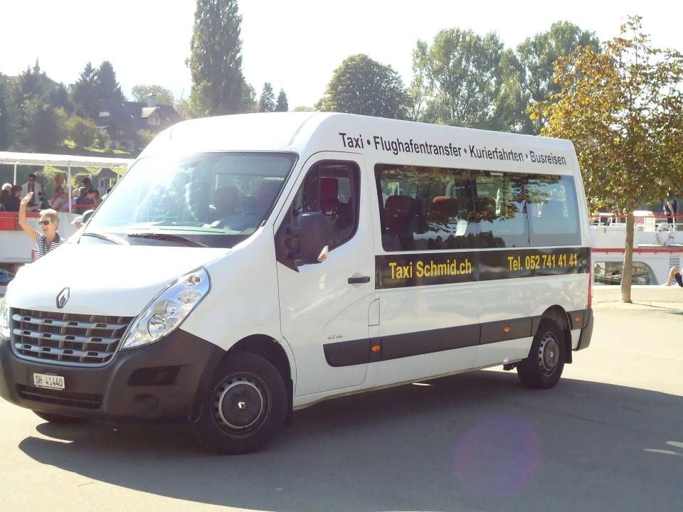Ein weisser Lieferwagen parkt in einer sonnigen Strasse, an der Seite steht „Taxi Schmid“.