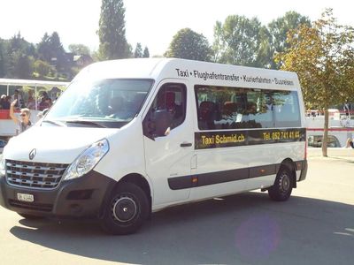 Ein weisser Lieferwagen parkt in einer sonnigen Strasse, an der Seite steht „Taxi Schmid“.