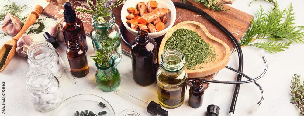 Verschiedene Kräuterheilmittel, Flaschen und getrocknete Zutaten auf einem weissen Tisch mit Blättern und Schalen.