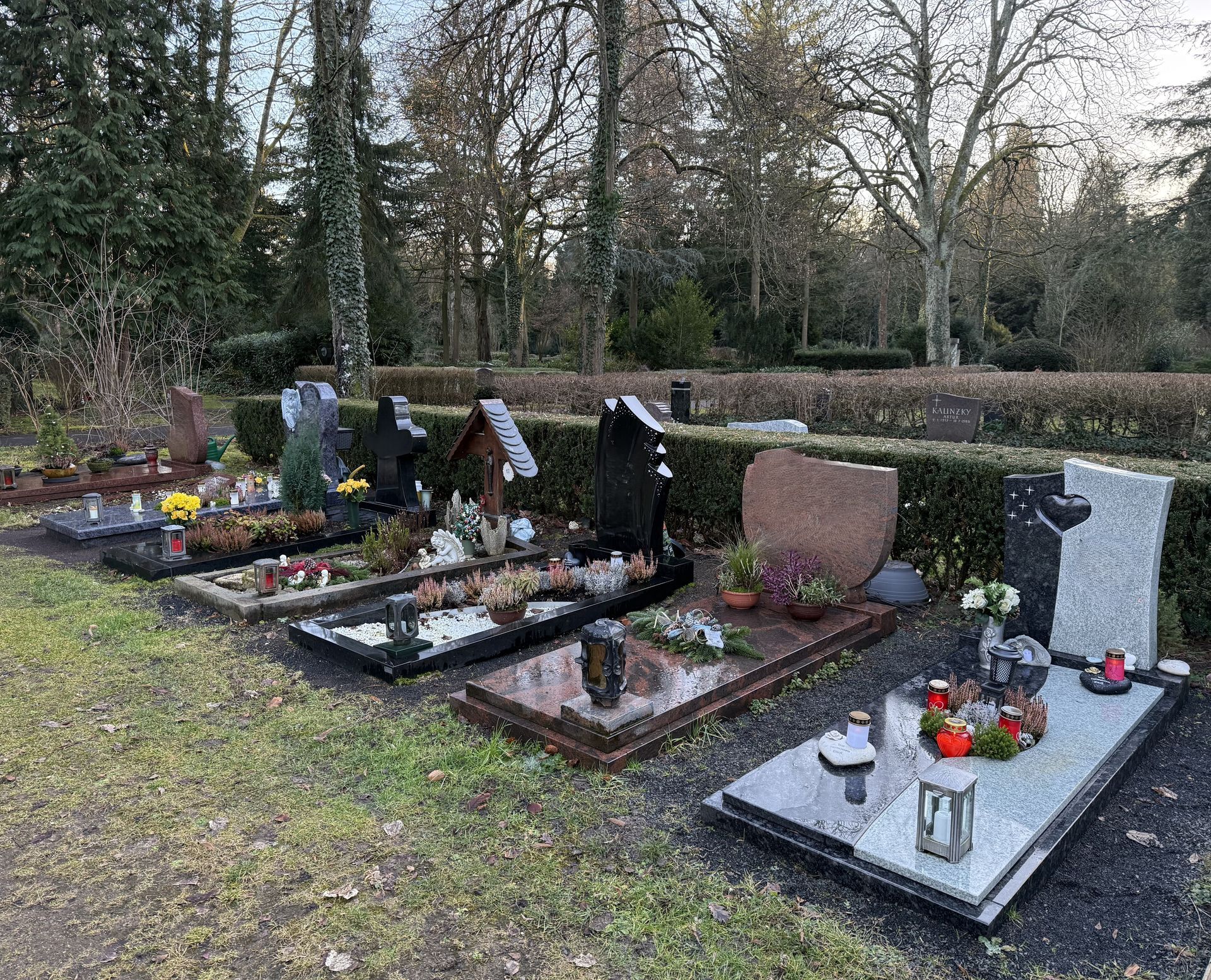 Mehrere Grabstätten mit Blumen und Kerzen auf einem von Hecken und Bäumen umgebenen Friedhof.