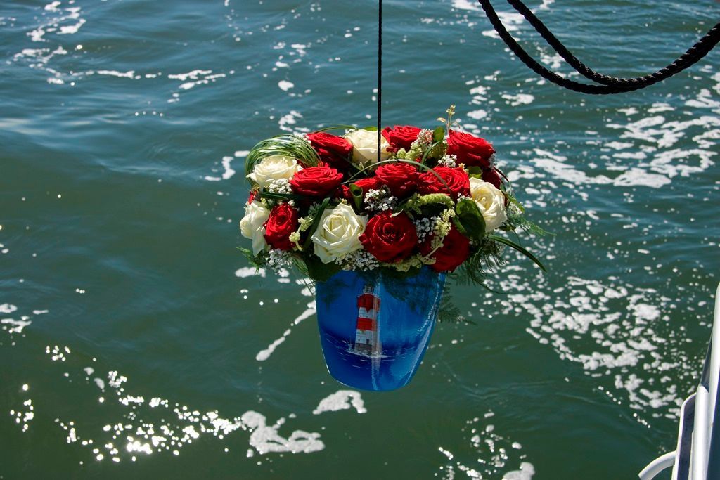 Eine blaue Urne mit roten und weißen Blumen hängt über glitzerndem grünen Wasser