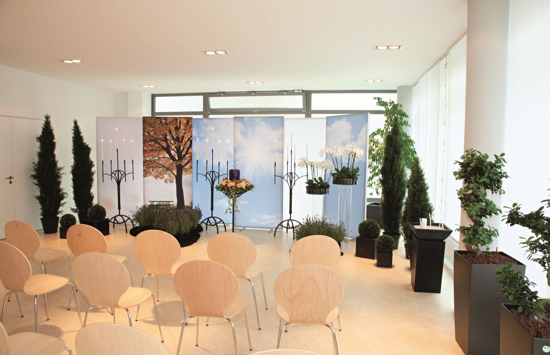 Heller Veranstaltungssaal mit Reihen weißer Stühle, Topfpflanzen und hängenden Blumenarrangements an großen Fenstern