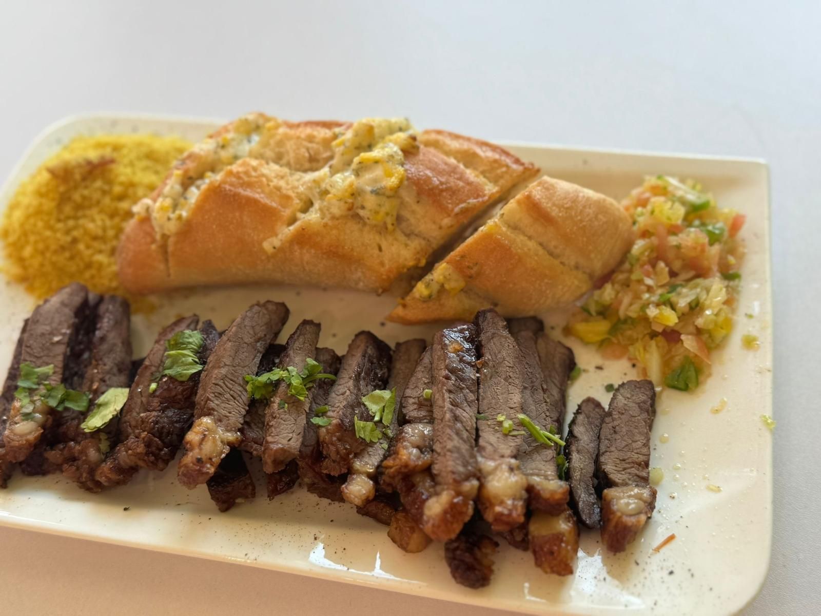 Filete en rodajas, pan con aderezo, guarnición amarilla y salsa en un plato rectangular blanco.