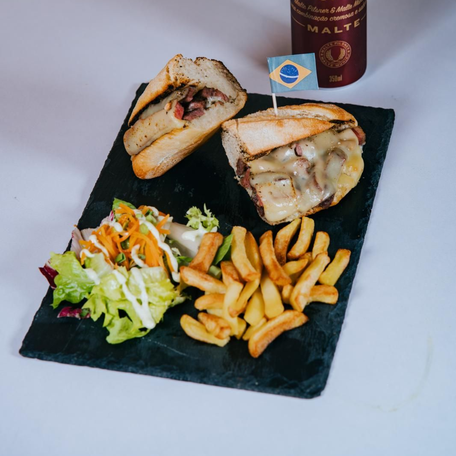 Sándwich con papas fritas, ensalada y una botella en un plato negro. El sándwich tiene una bandera de Brasil.