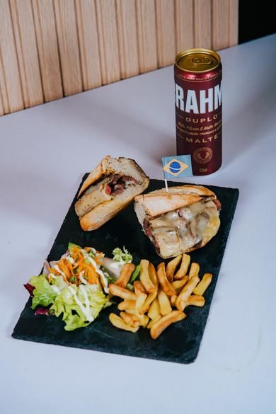 Un sándwich partido por la mitad con queso fundido, una ensalada y patatas fritas sobre una tabla de pizarra, con una lata de cerveza Brahma detrás.