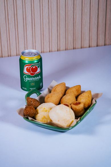 Una lata verde de refresco Guaraná Antarctica junto a una pequeña cesta verde que contiene aperitivos brasileños fritos y pão de queijo.
