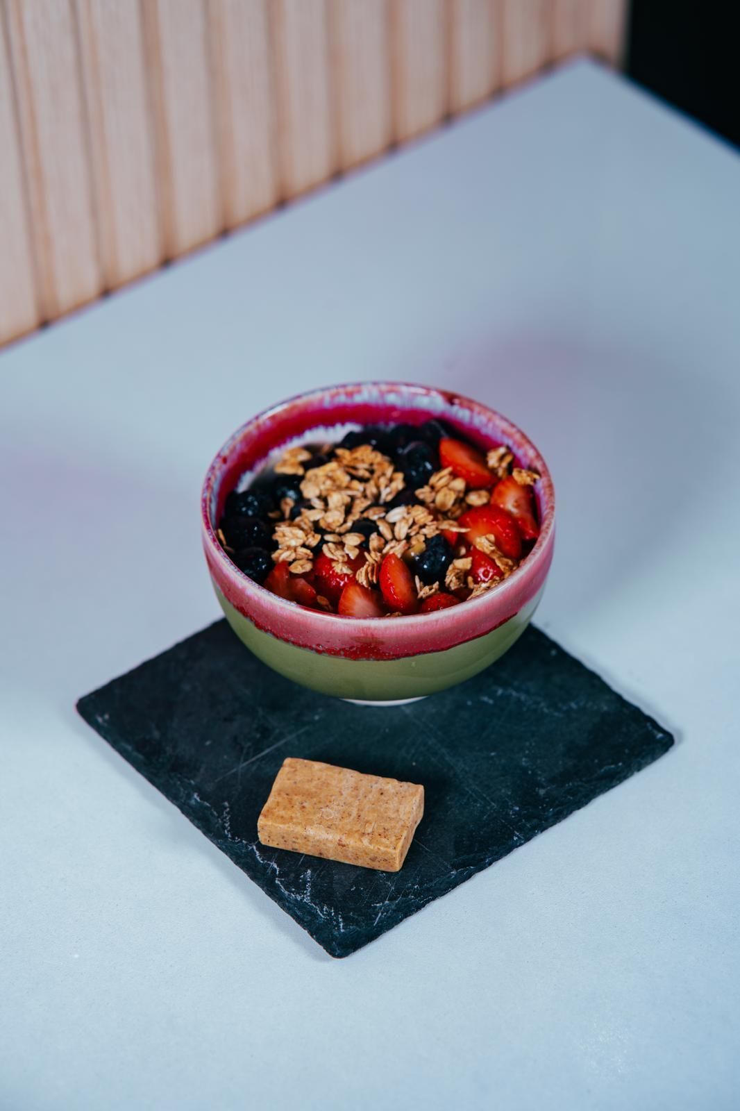 Tazón de bayas y granola sobre cuadrado negro, con galleta, sobre superficie blanca.