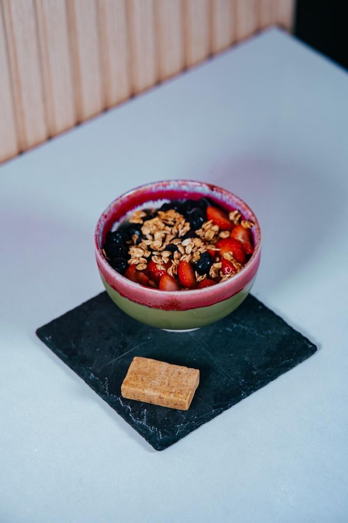 Un bol de cerámica rosa y verde con yogur, frutos rojos y granola, servido con una galleta en una bandeja de pizarra negra.