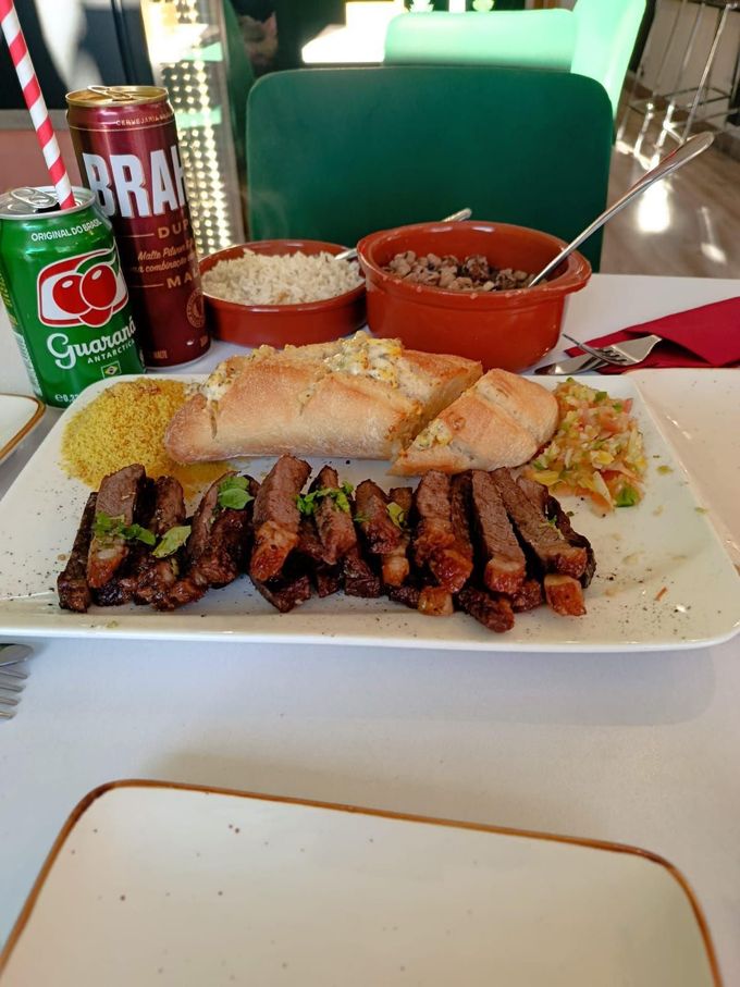 Un plato típico de restaurante que incluye filete de picanha en rodajas, pan tostado, farofa y tazones de arroz con frijoles, acompañado de refrescos.
