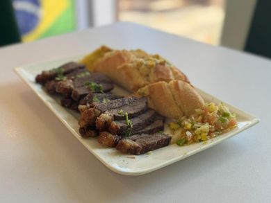 Un plato de carne de res en rodajas, pan de ajo y salsa, con una bandera brasileña borrosa al fondo.