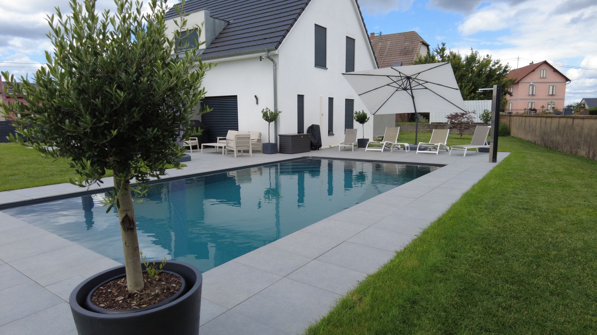 Piscine en béton avec arbre en pot et vue sur une terrasse avec un grand parasol.