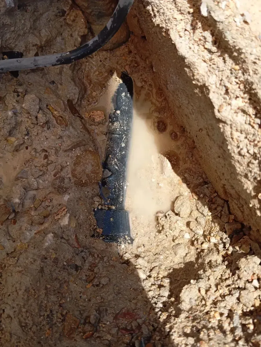 Vista de cerca de una tubería negra reventada que gotea agua en una zanja de tierra.