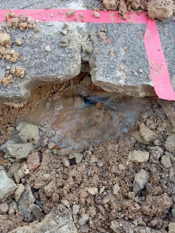 Una tubería reventada arroja agua a un agujero fangoso excavado en el suelo, marcado con tiras de cinta roja de precaución.