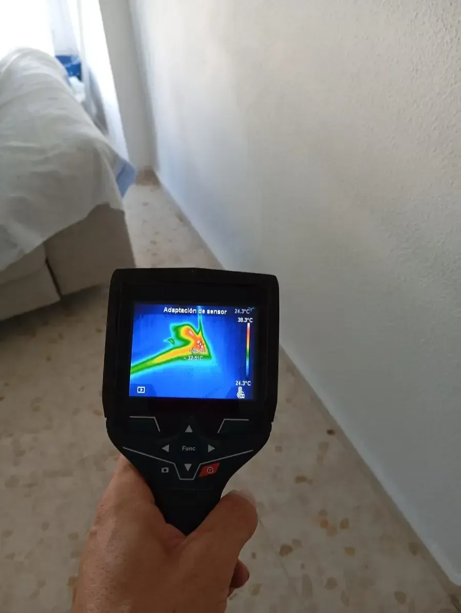 Una mano sostiene una cámara termográfica apuntando a una pared, mostrando una señal de calor localizada en su pantalla.