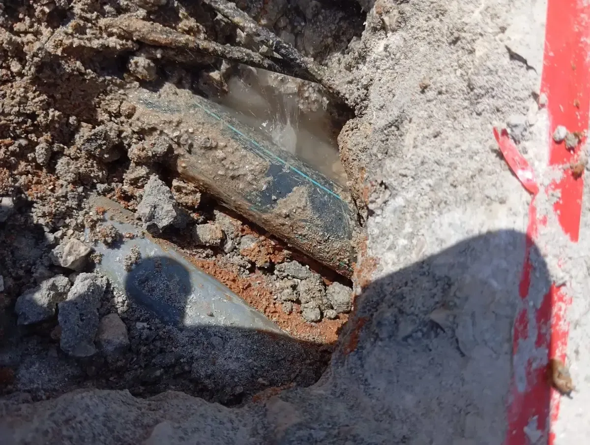 Dos tuberías grises yacen expuestas en una zanja fangosa y llena de tierra, junto a una sección de hormigón rojo.