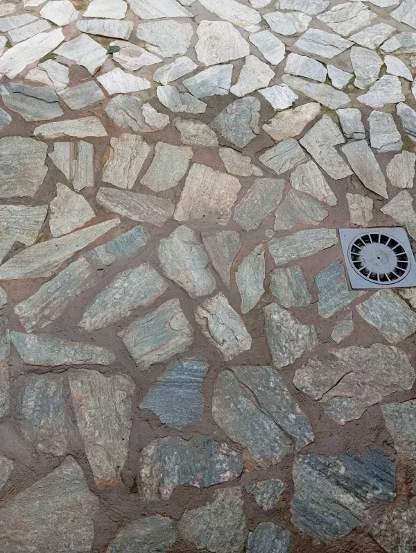 Patio pavimentado con piedras grises irregulares, lechada marrón y una rejilla de drenaje metálica cuadrada.