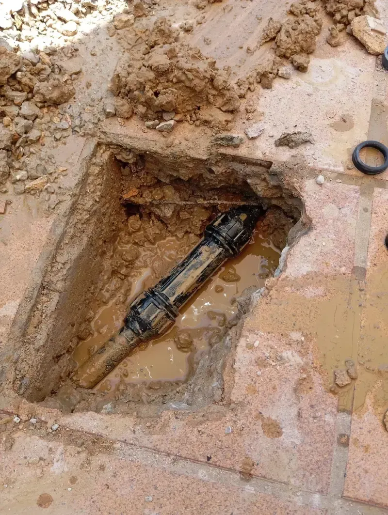Un conector de tubería de plástico negro instalado en un agujero excavado y fangoso en una superficie pavimentada.